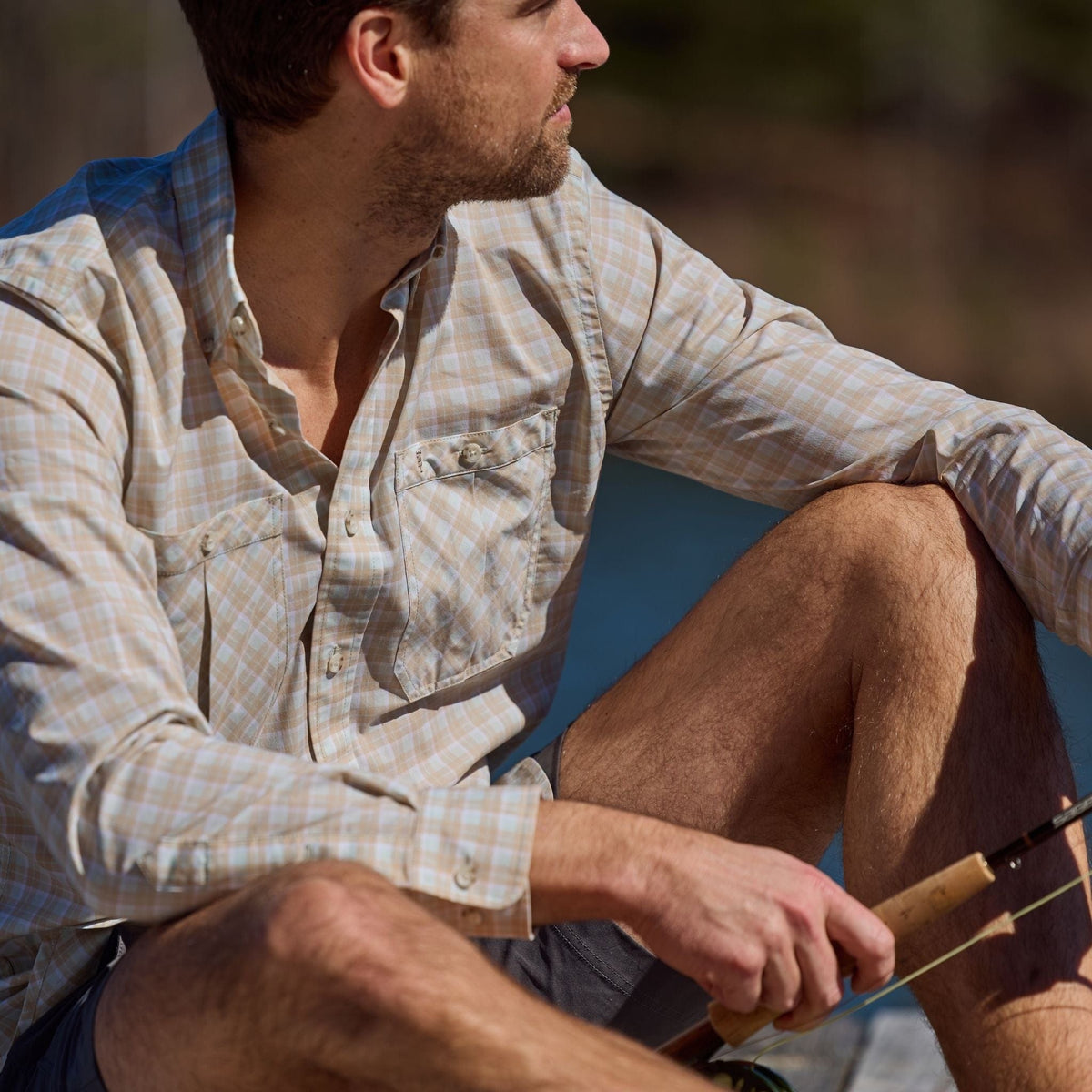 Man in a Tom Beckbe Tidewater Shirt and shorts sits by the water with a fishing rod, looking away from the camera.