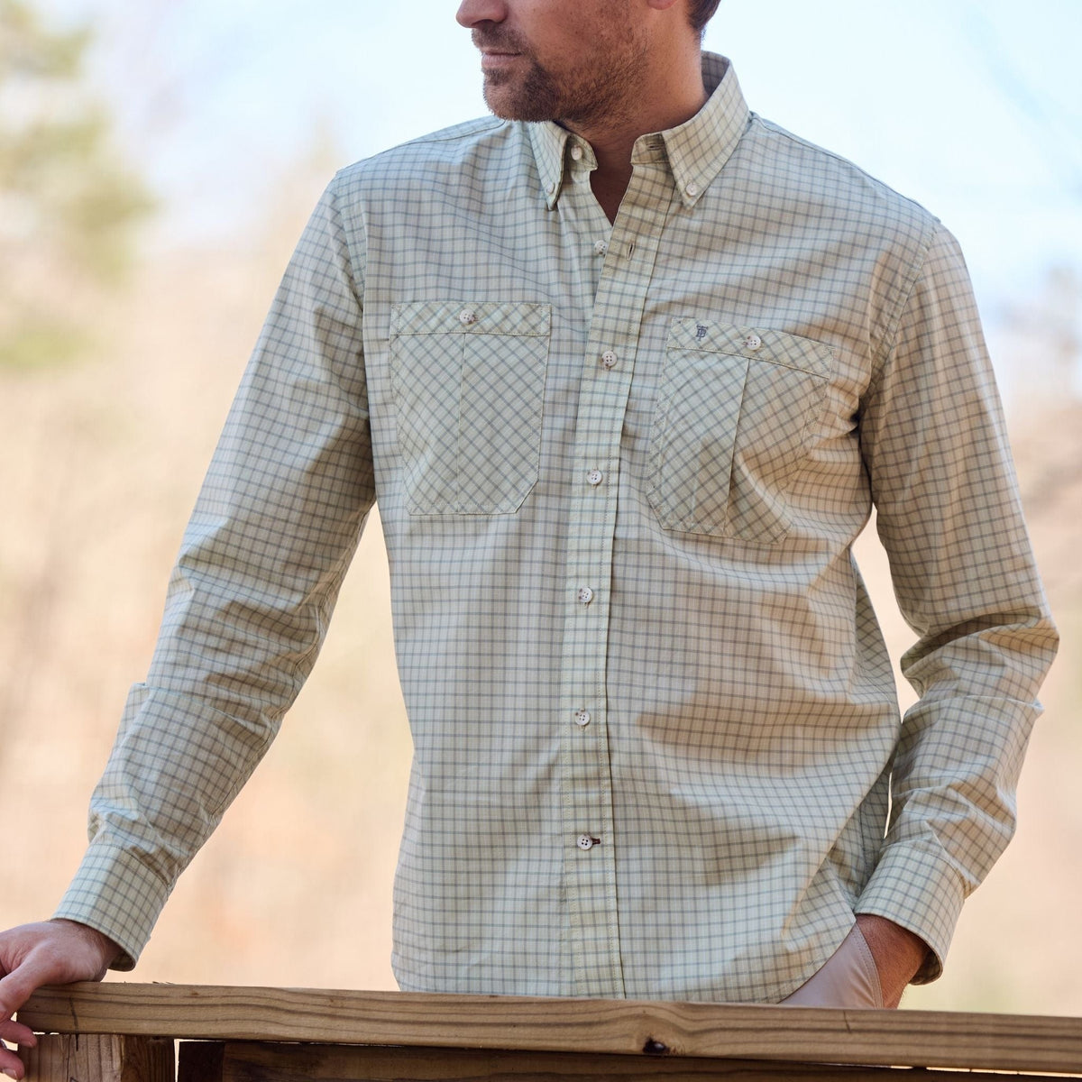 A man in a Tom Beckbe Tidewater Shirt leans on a railing; blurred trees suggest adventure behind UPF 30+ protection.