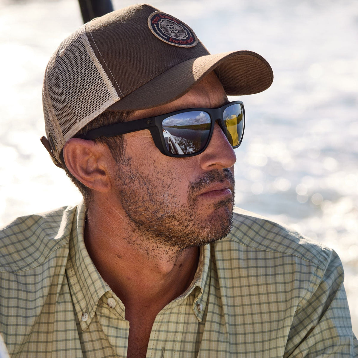 A man in a cap and sunglasses calmly gazes over the water in a Tom Beckbe Tidewater Shirt with UPF 30+ coverage.