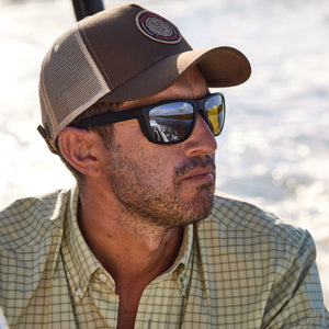 A man in a cap and sunglasses calmly gazes over the water in a Tom Beckbe Tidewater Shirt with UPF 30+ coverage.