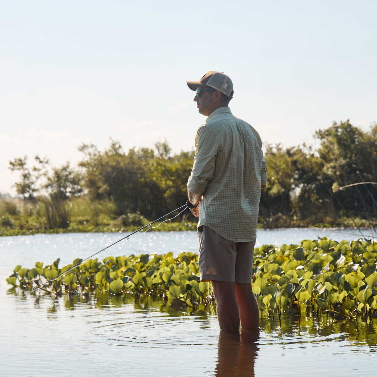 A man in a Tom Beckbe Tidewater Shirt stands knee-deep fishing among plants beneath a clear sky.