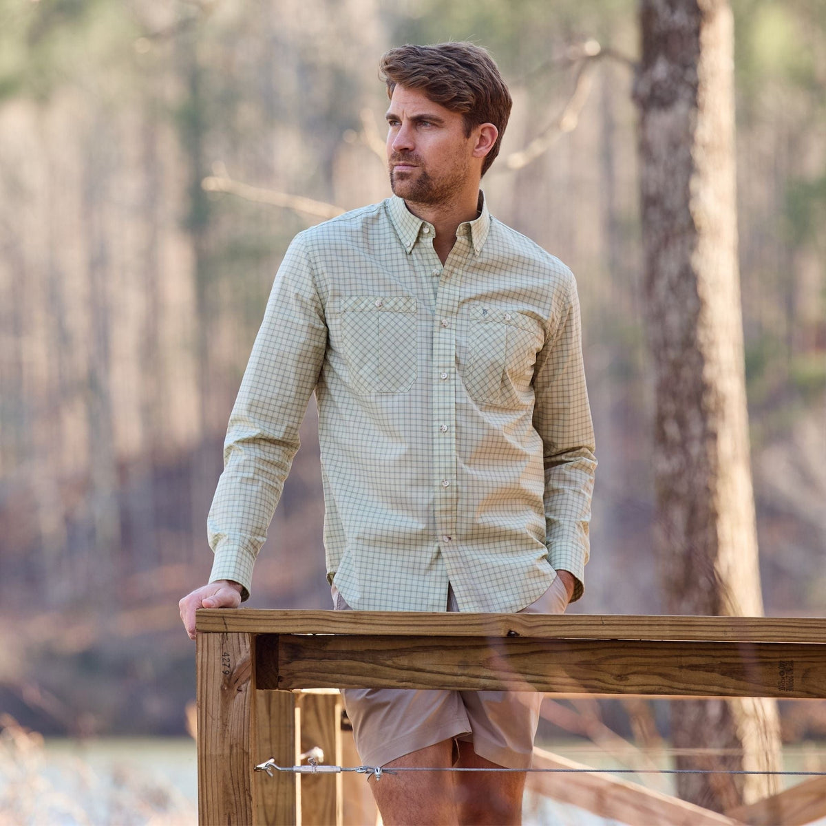 Man in a Tom Beckbe Tidewater Shirt and khaki shorts stands on a deck, gazing into the distance with trees behind.