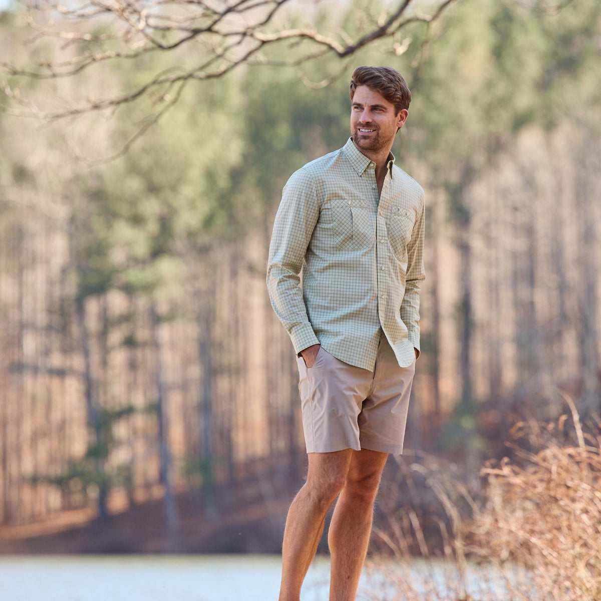 Man in a Tom Beckbe Tidewater Shirt stands by a lake with trees behind, gazing into the distance and smiling.