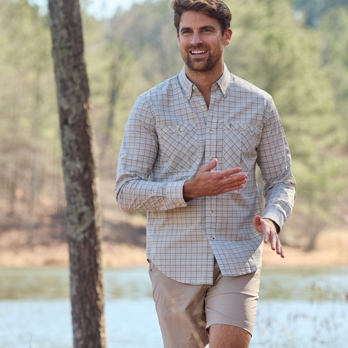 A bearded man in a Tom Beckbe Tidewater Shirt and shorts strolls outdoors, with trees and a lake behind him.