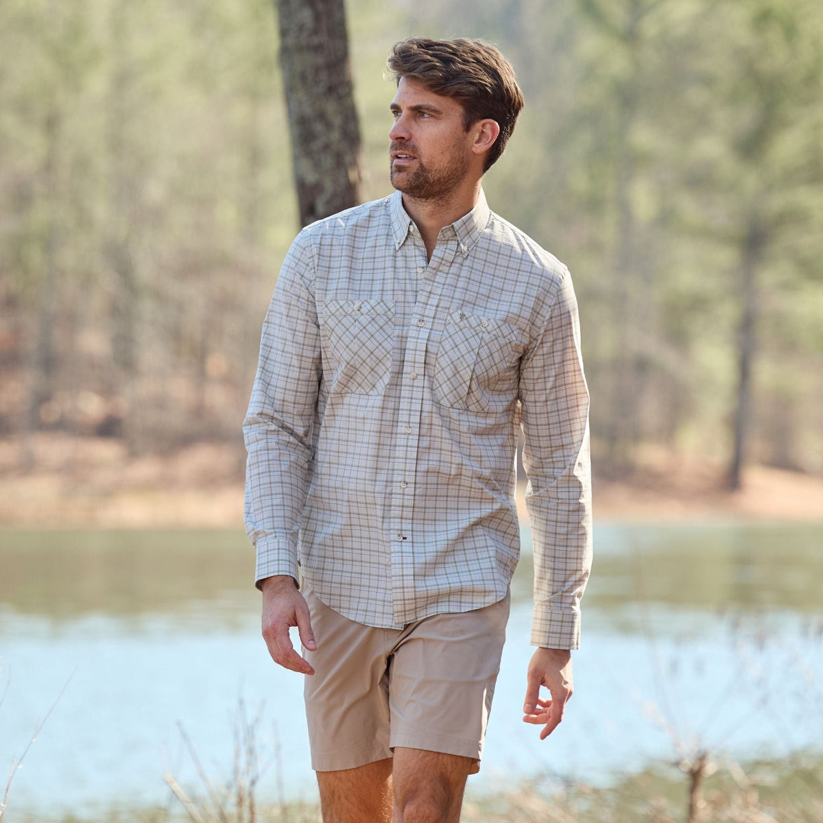 A man in a Tom Beckbe Tidewater Shirt and shorts strolls by a lake, surrounded by trees.