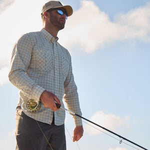 Man in a hat and sunglasses with a rod, wearing a Tom Beckbe Tidewater Shirt against a blue sky backdrop.