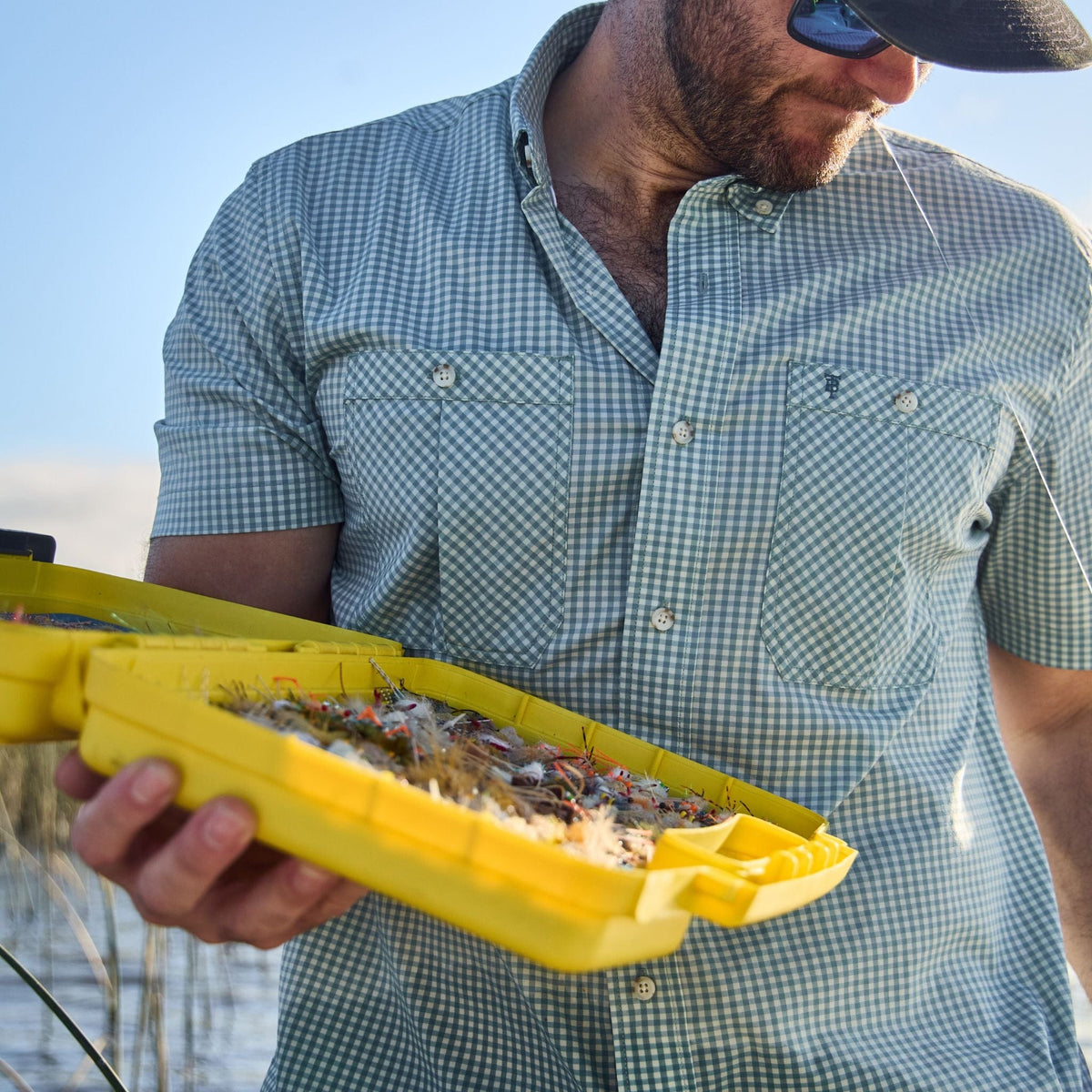 Man in a Tidewater Shirt by Tom Beckbe holding a yellow tackle box, enjoying a sunny day outdoors.