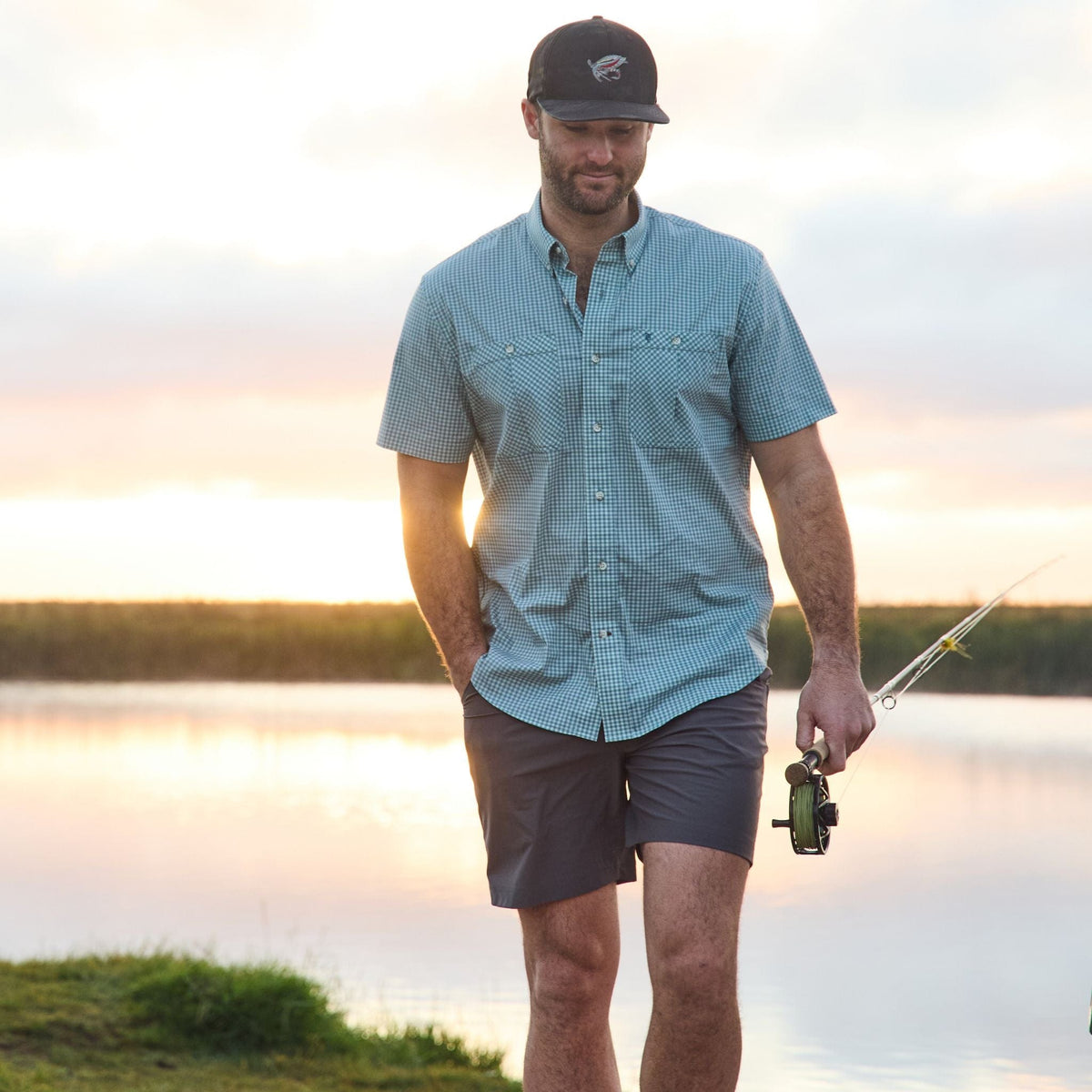 A man in a Tidewater Shirt by Tom Beckbe strolls by the lake at sunset with a rod.