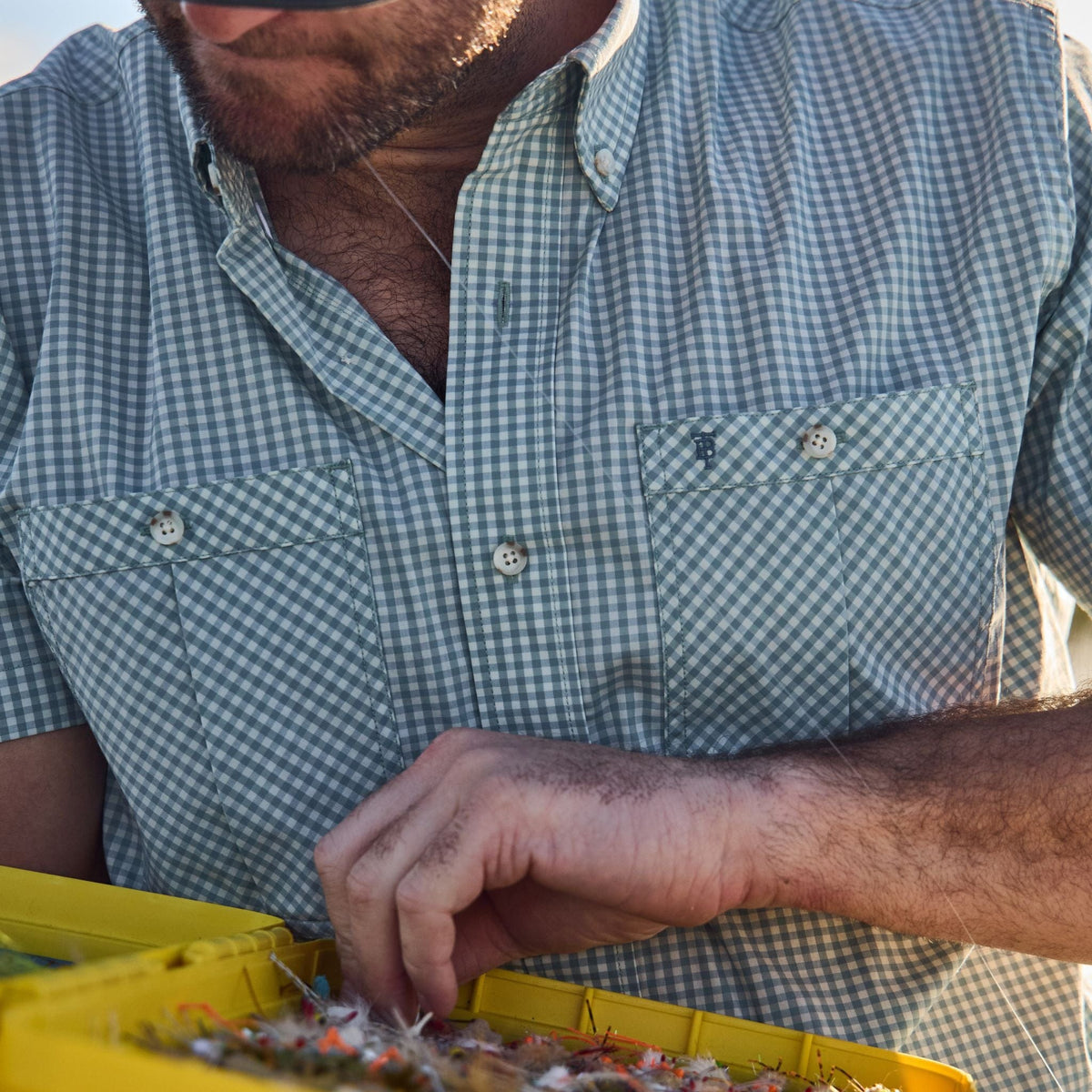 A person in a Tom Beckbe Tidewater Shirt inspects fishing flies in a yellow tackle box.