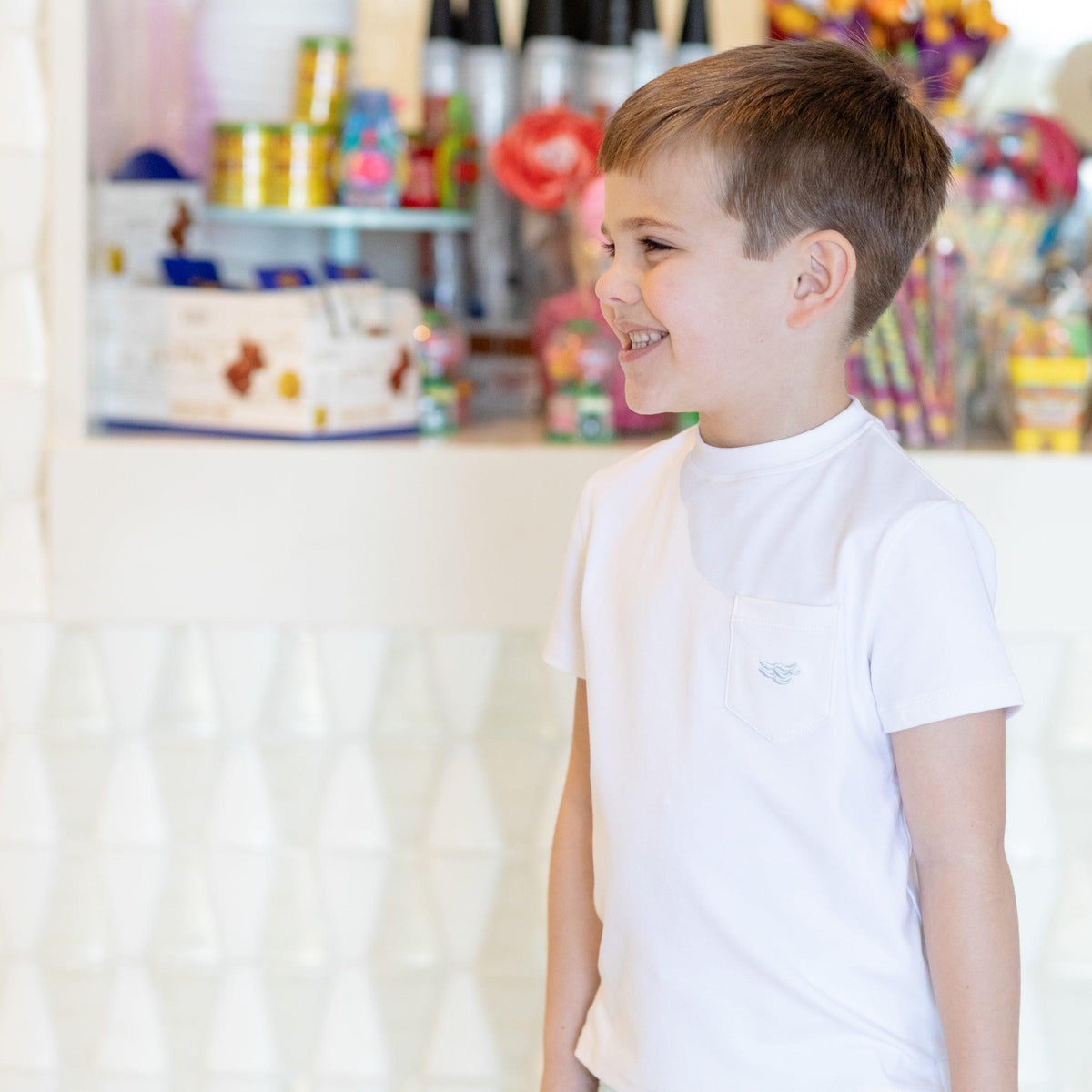 Right-side view of Tucker Tee in Classic White showcasing short sleeves and chest pocket on a smiling boy in a candy-shop background