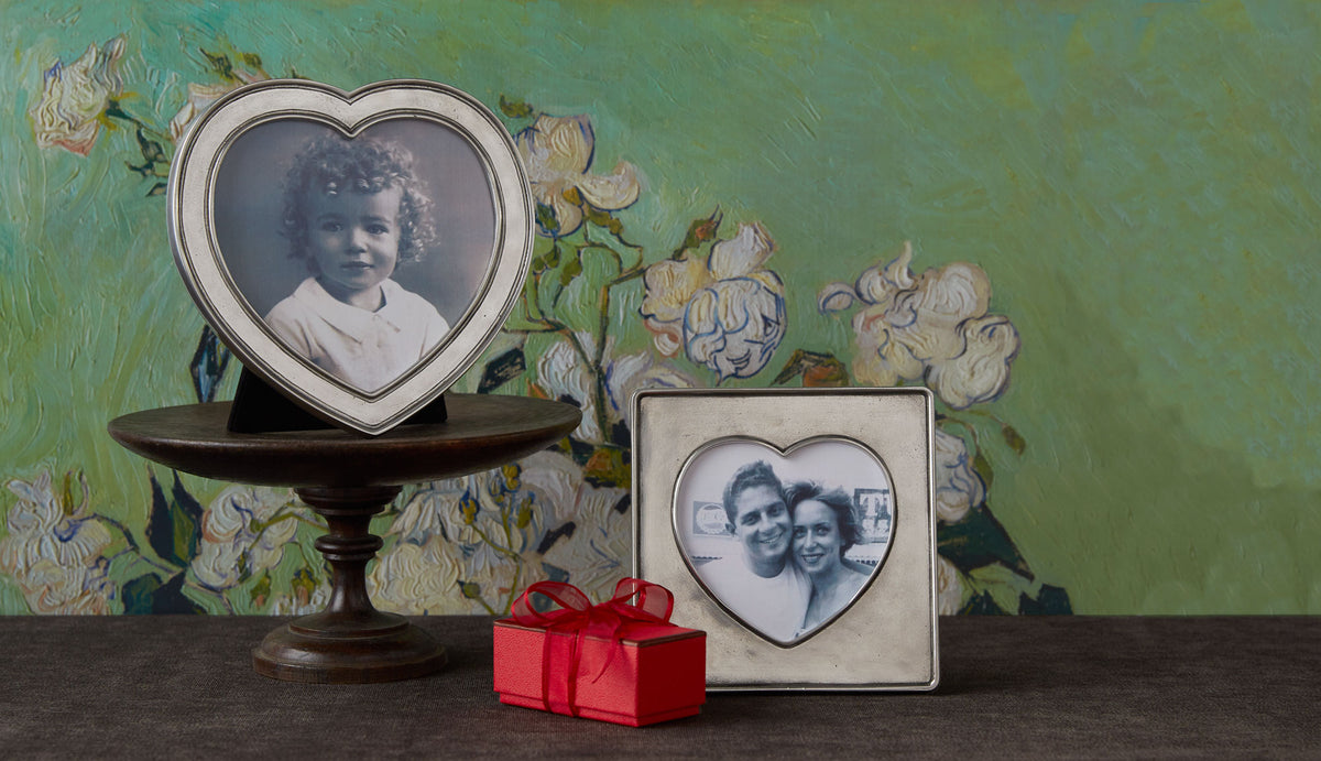 Front view of pewter heart frame on wood pedestal showing heart-shaped opening and soft pewter luster with small red gift box and painted floral backdrop
