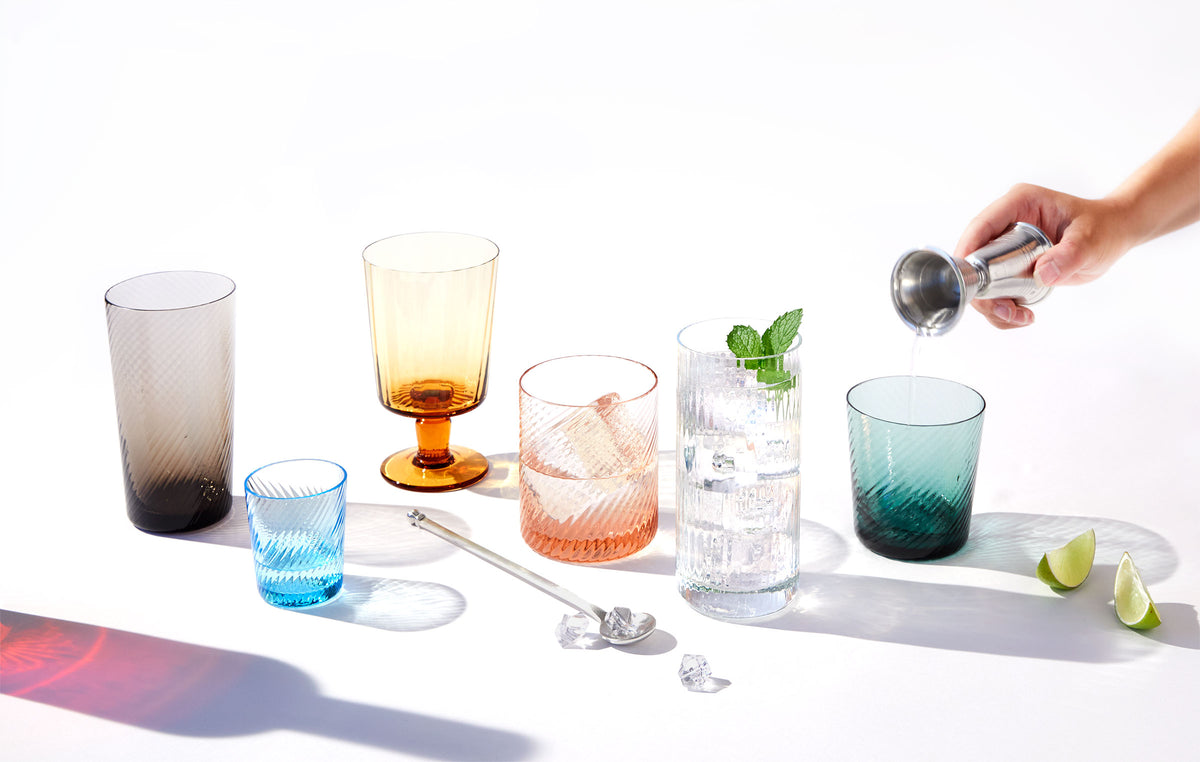 Assorted Bembo Twist glasses in jewel tones with a hand pouring liquid into one glass on a white surface with lime wedges and a bar spoon