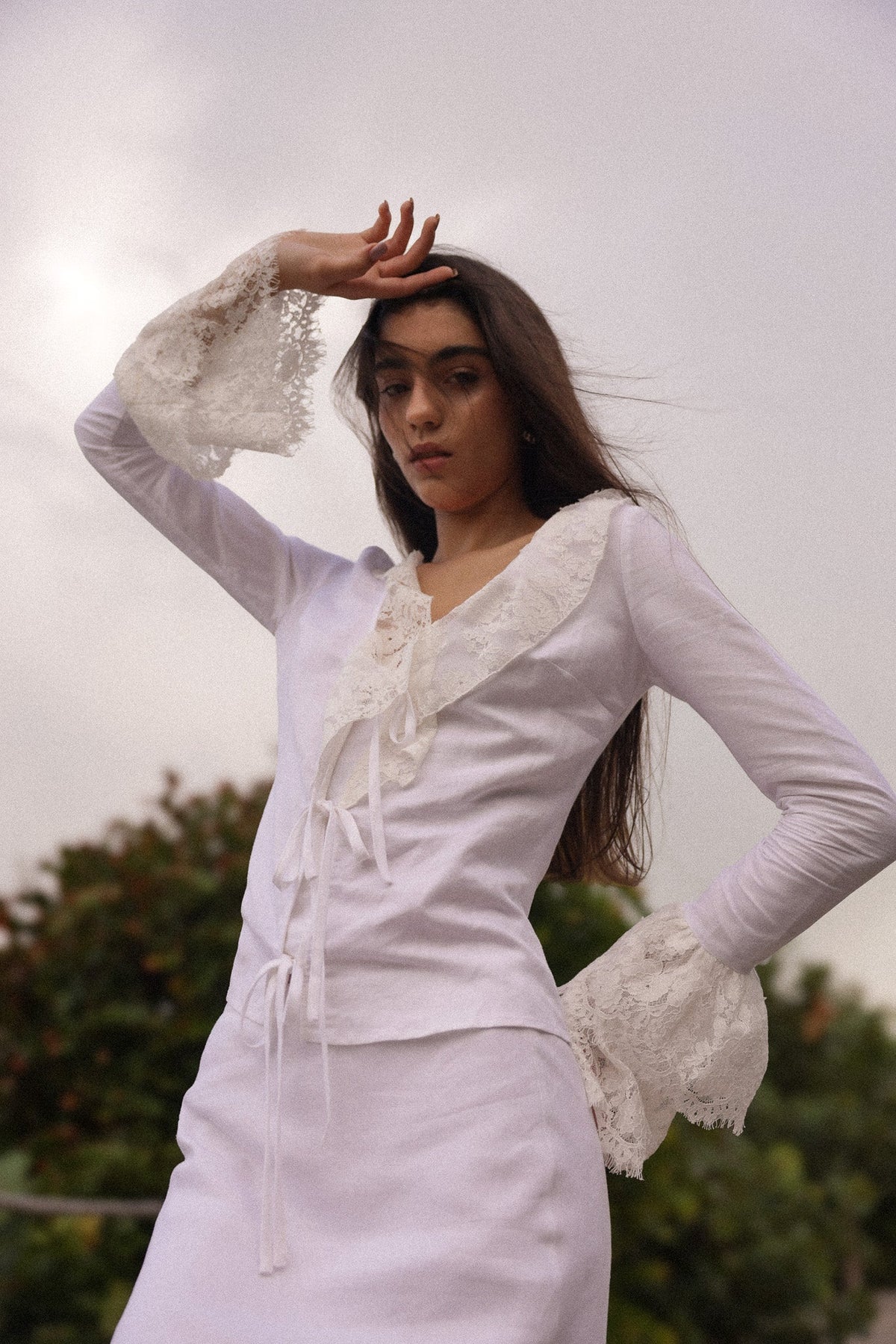 Model wearing a fitted white linen tie-front top with lace trim and dramatic bell sleeves, styled for a romantic summer evening look by Sister New York.