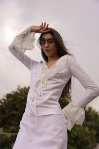 Model wearing a fitted white linen tie-front top with lace trim and dramatic bell sleeves, styled for a romantic summer evening look by Sister New York.