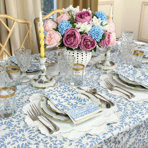 Elegant table setting with floral centerpiece, blue leaves patterned tablecloth, white vegan leather embroidered placemats and decorative glasses.