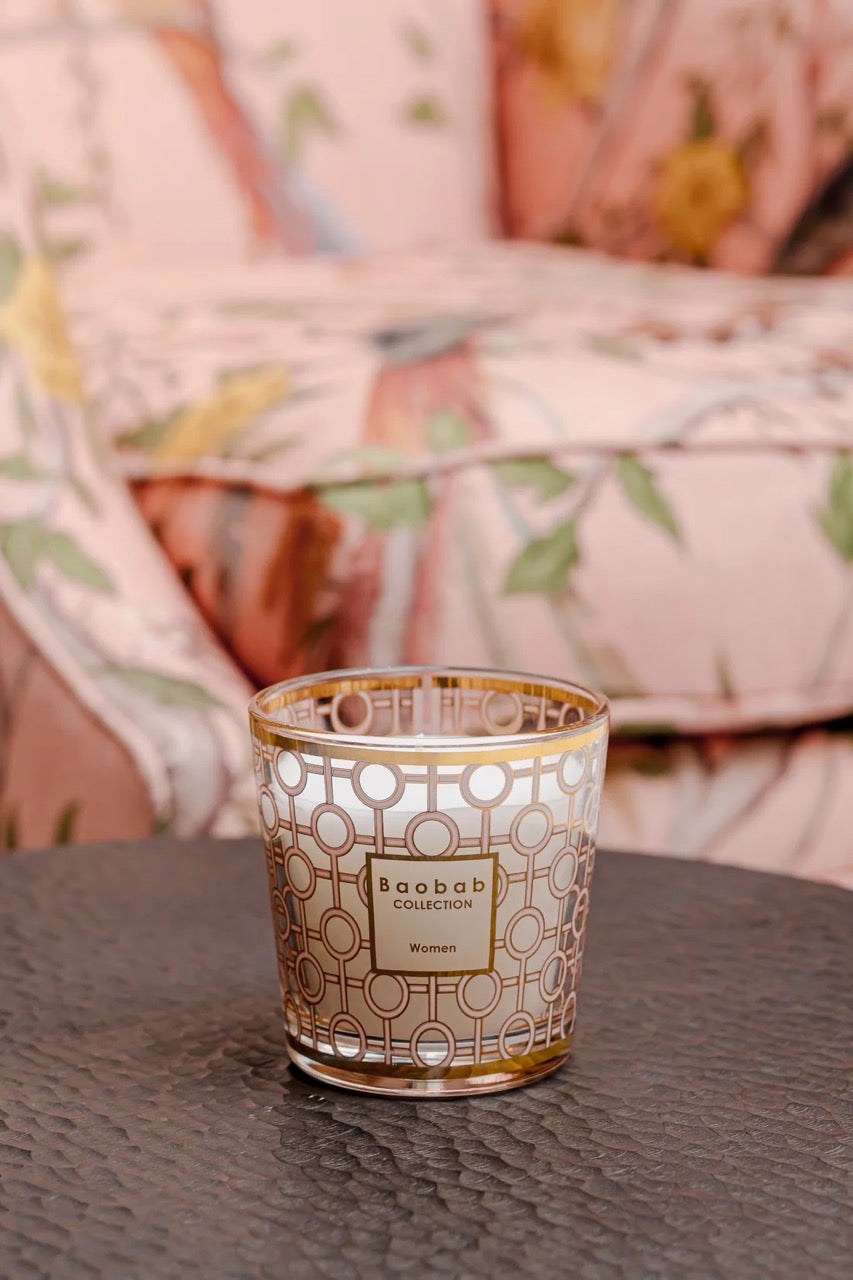 Front view of Baobab Women candle in pink glass with gold geometric pattern on tabletop and blurred floral sofa