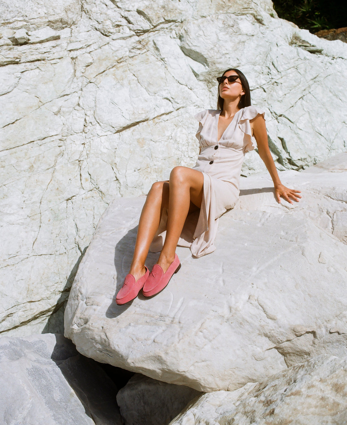 Woman seated on sunlit white rocks wearing Begonia pink suede Milano loafers and a beige ruffled dress
