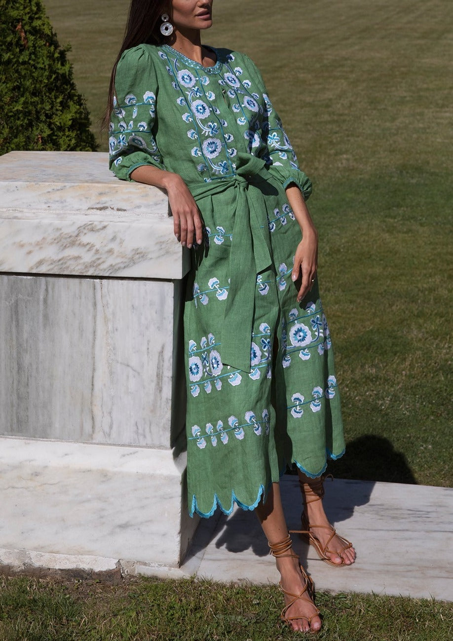Three-quarter view of Emerald Lucia dress showcasing blue floral embroidery, scalloped hem and tie belt by a marble ledge