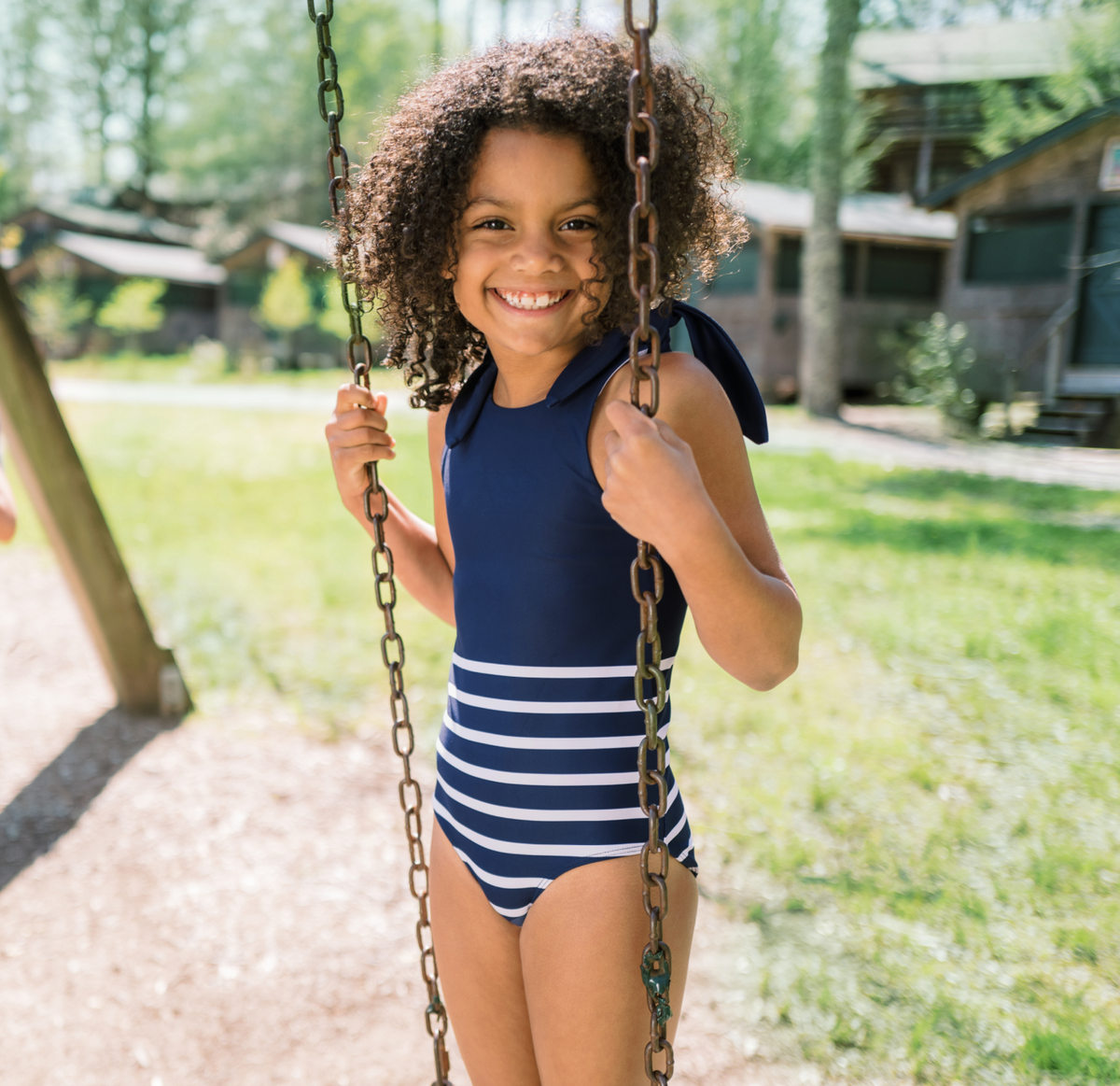 Front view of Girls Navy Breton Stripe Tie-Knot One Piece showing navy Breton stripes and shoulder ties on a swing outdoors
