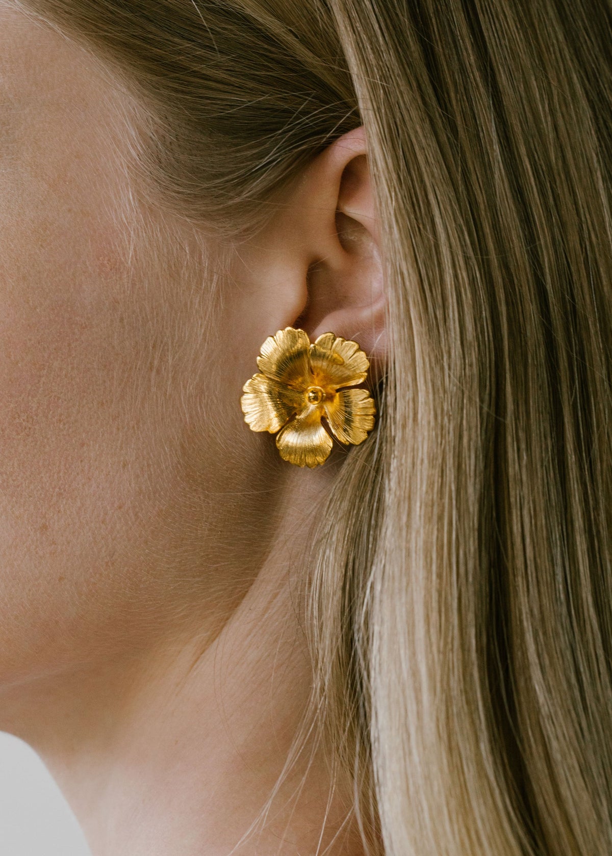Close-up of Rowena Earrings showcasing a light antique gold-tone flower design on a model’s ear with blonde hair