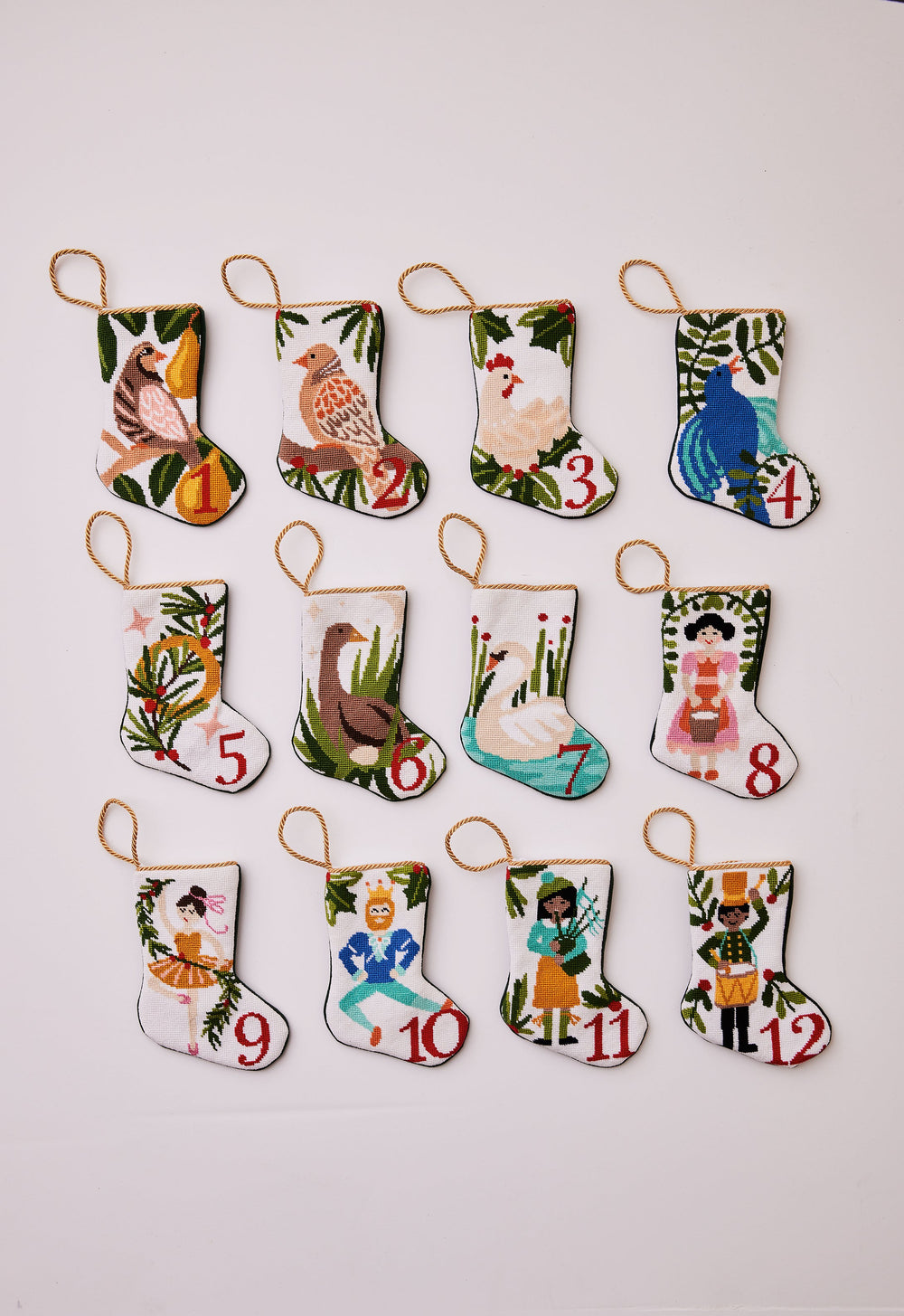 Overhead view of 12 Days of Christmas bauble stocking set showing 12 numbered needlepoint mini stockings with gold hanging loops on pale background