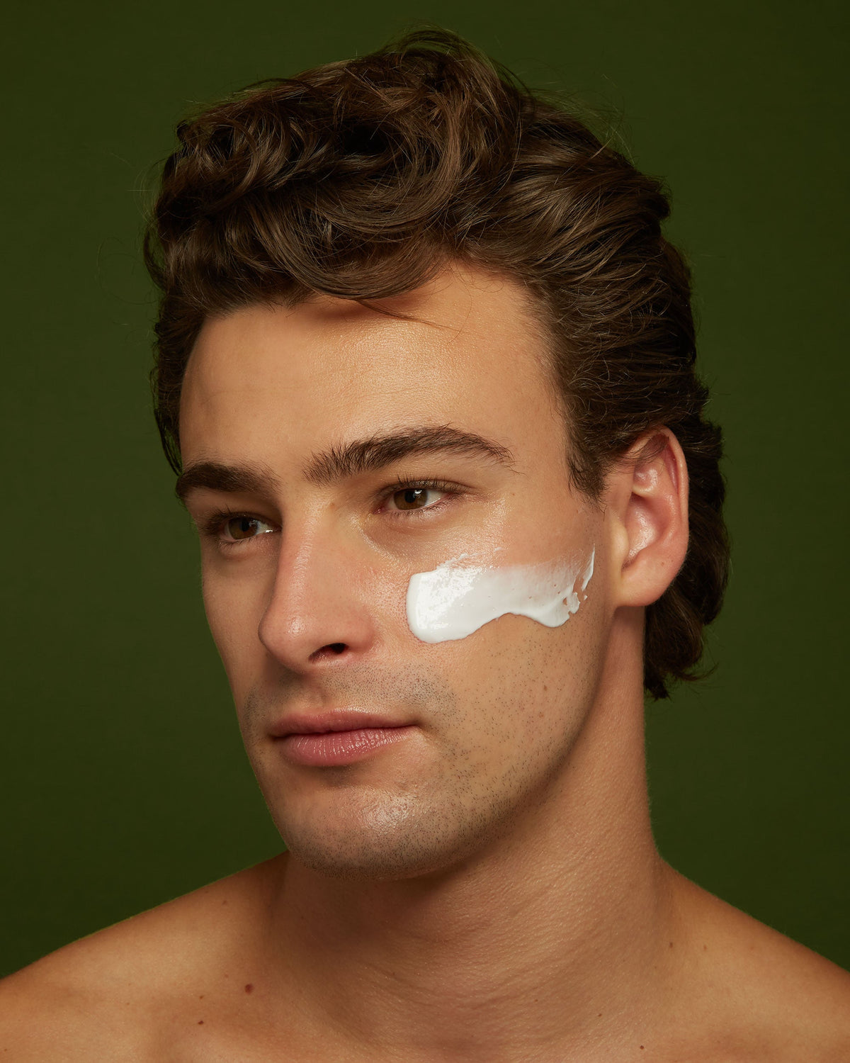 Young man with a swipe of moisturizer on his right cheek, close-up portrait on an olive green background