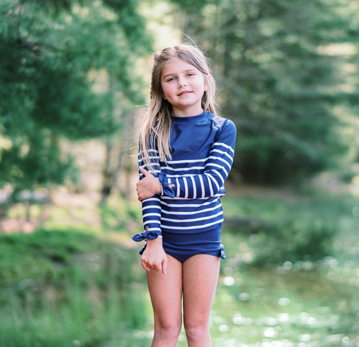Front view of Girls Navy Breton Stripe Rashguard Set showcasing striped long sleeves and bow cuffs with a blurred wooded background