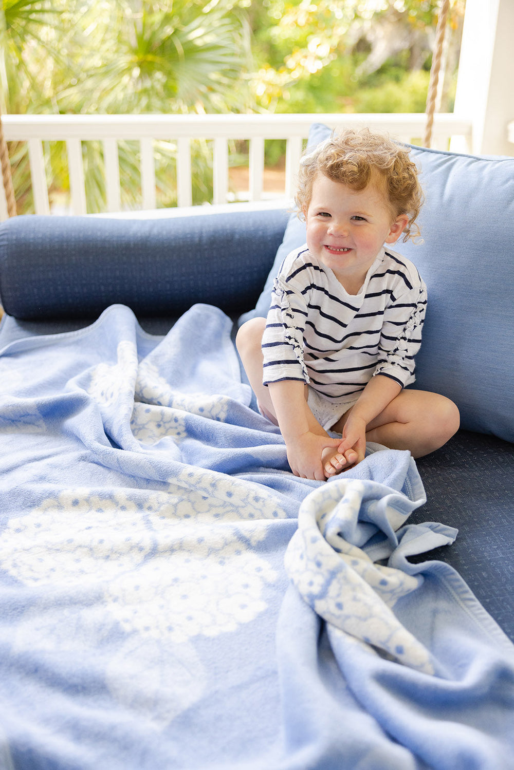 Angled view of Hydrangeas Bluebell blanket showing blue hydrangea jacquard pattern draped on a porch seat with a toddler