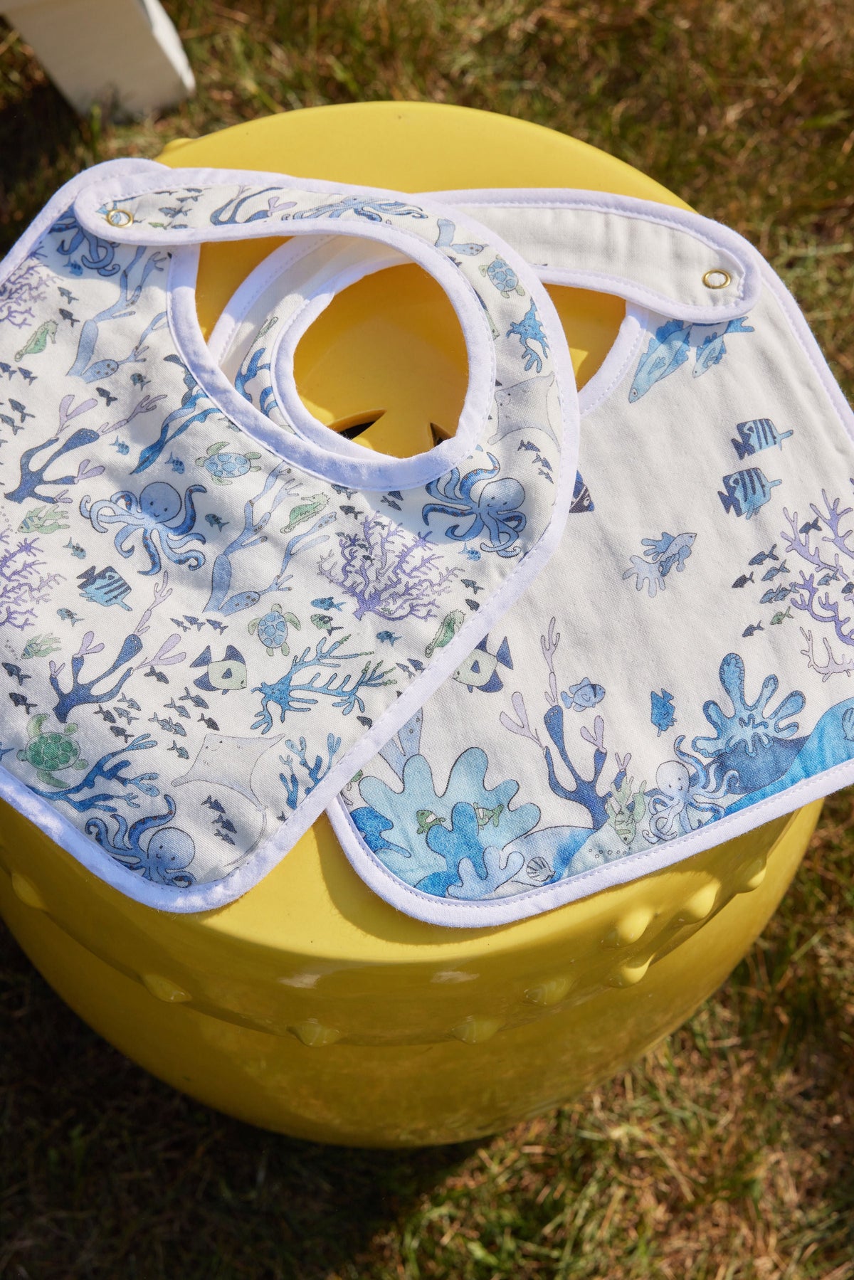 Flat lay of two Coral Reef bibs showcasing blue ocean print, white trim, and snap closures on a yellow stool