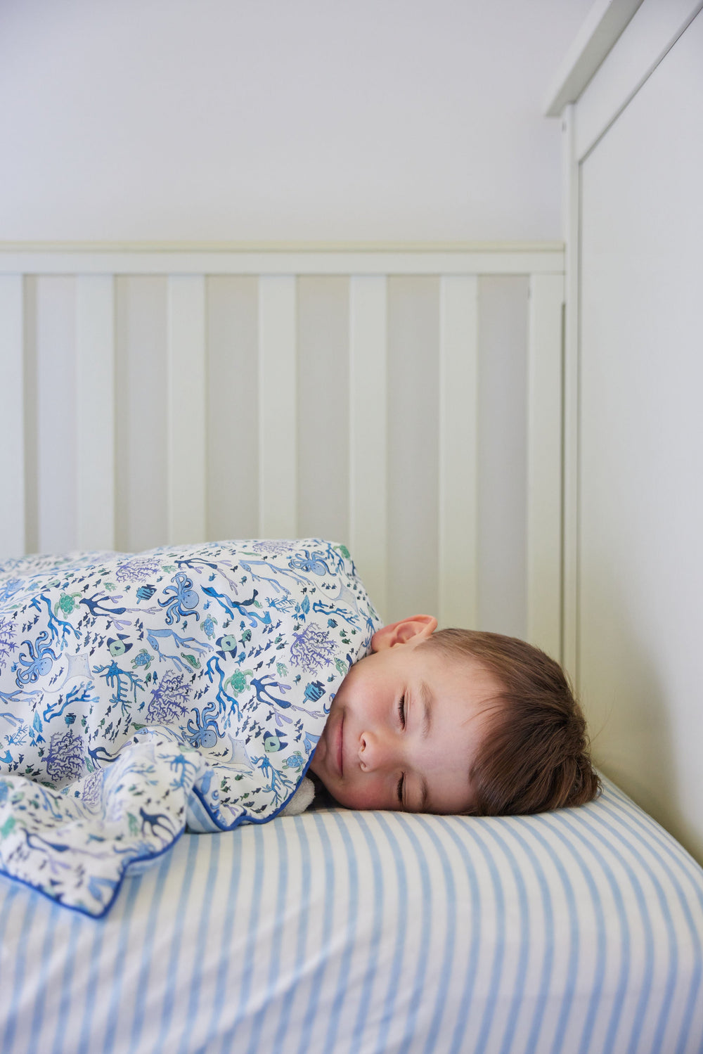 Toddler sleeping on a blue and white striped crib sheet in a white slatted crib, covered by an ocean-print blanket