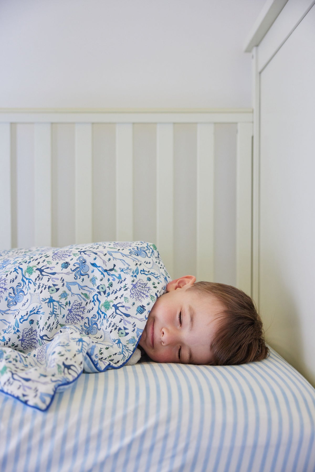 Toddler sleeping on a blue and white striped crib sheet in a white slatted crib, covered by an ocean-print blanket