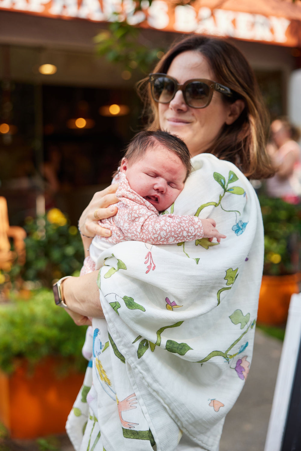 Woman holding a sleeping newborn wrapped in a white Tropical Chinoiserie shawl blanket with green vine and pastel floral and bird print outdoors