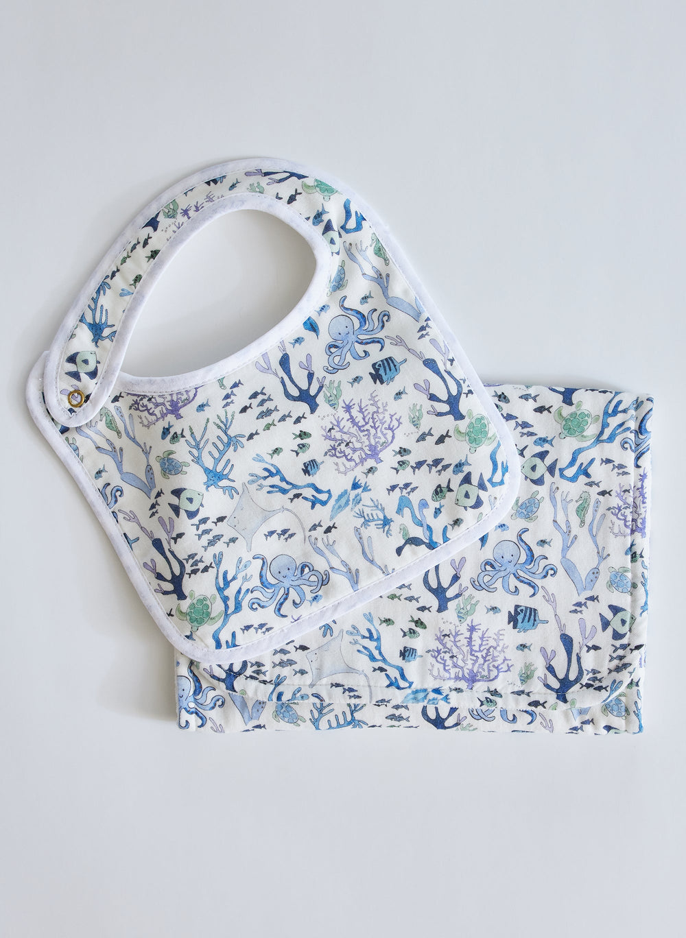 Flat lay Coral Reef bib and burp cloth set with blue-green sea life print on white muslin, white trim and snap closure