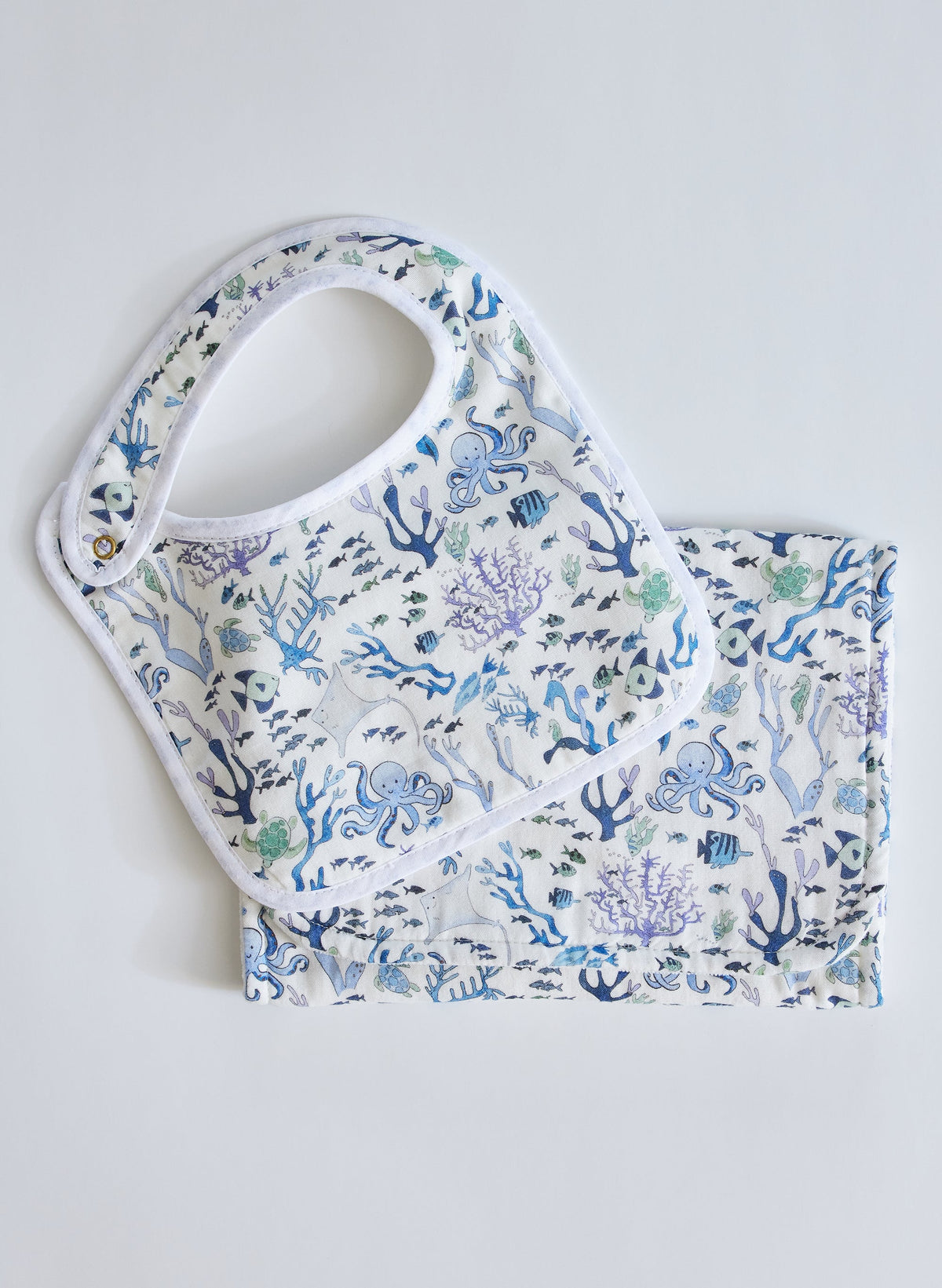 Flat lay Coral Reef bib and burp cloth set with blue-green sea life print on white muslin, white trim and snap closure
