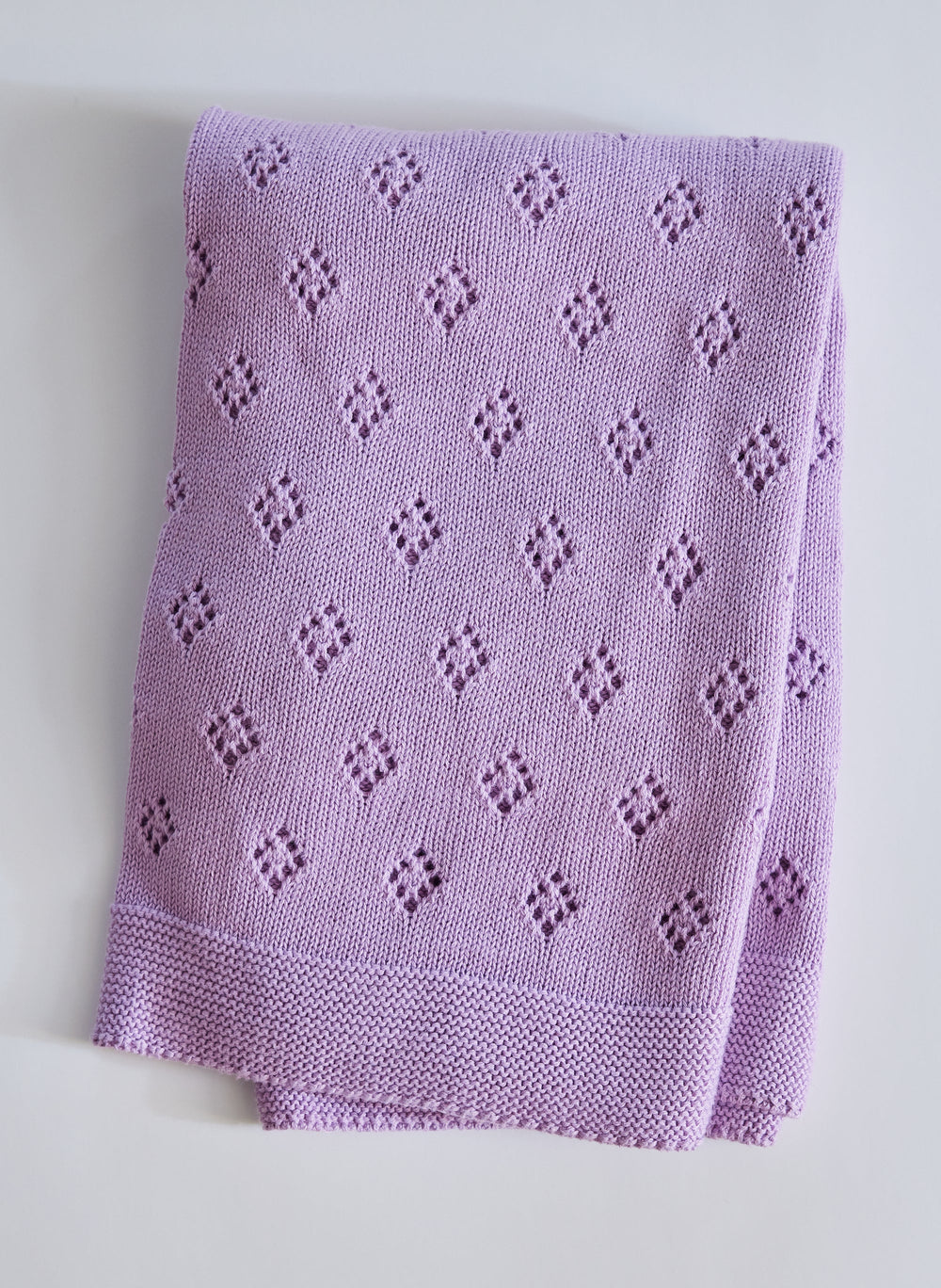 Folded view of The Wander Baby Blanket in lavender with diamond eyelet pattern and garter stitch border on gray background