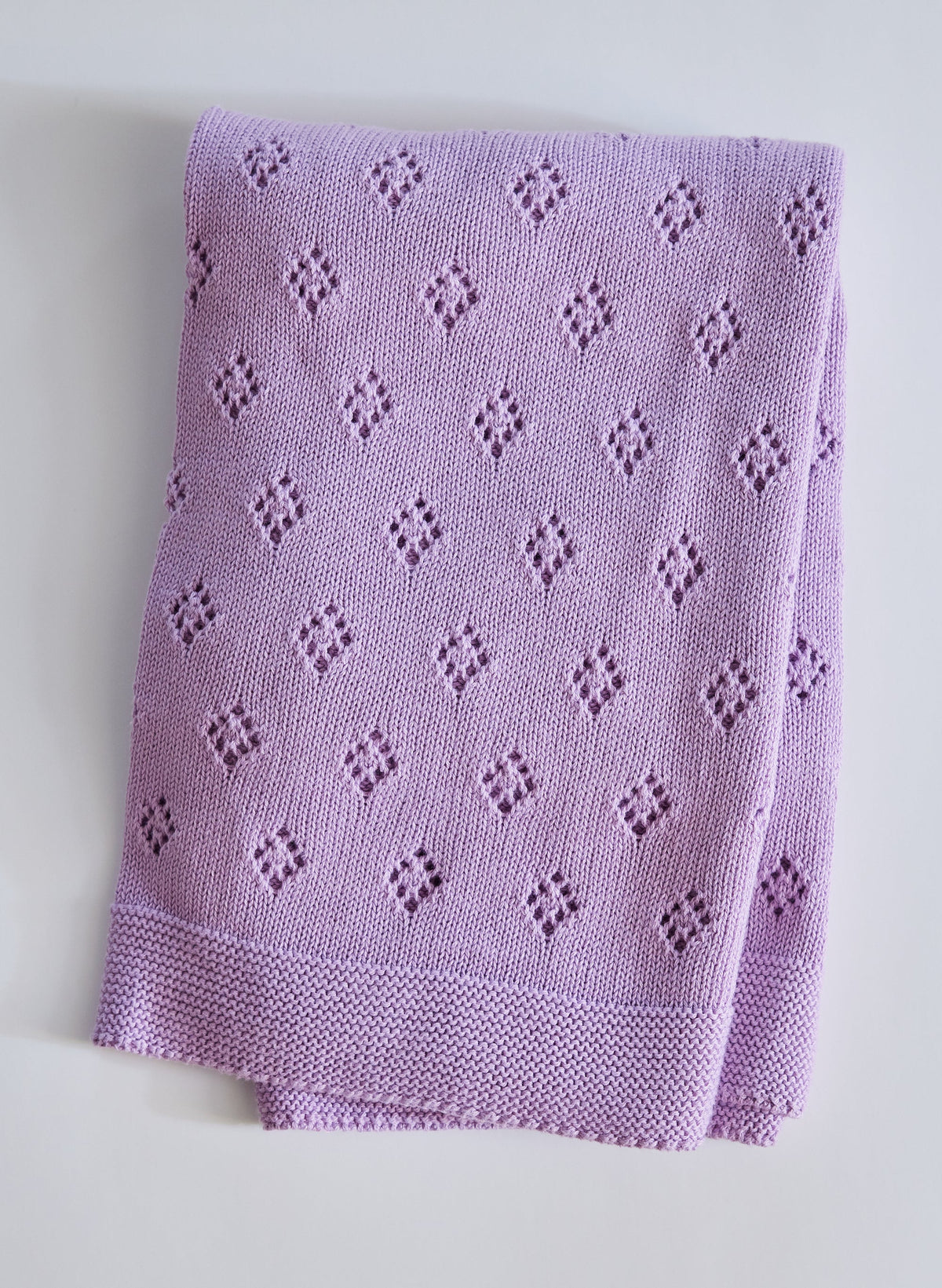 Folded view of The Wander Baby Blanket in lavender with diamond eyelet pattern and garter stitch border on gray background