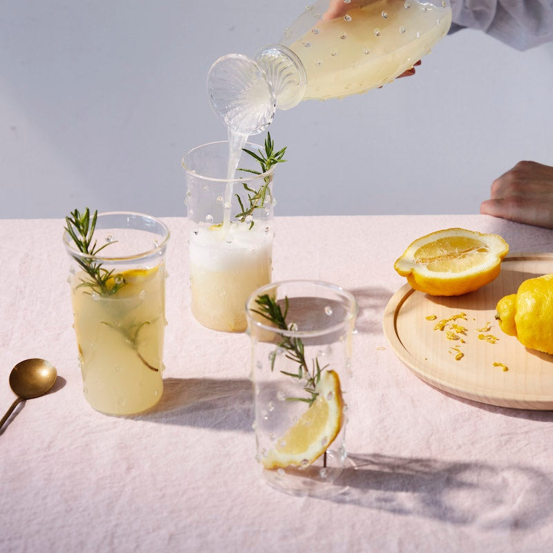 Three clear Pom highball glasses with raised bobble dots, one being filled from a matching carafe on a pink tablecloth with lemon halves and rosemary garnish