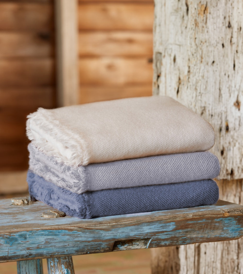 Stacked view of Noe Cashmere Throw in Arctic Blue showing fringed edge and soft herringbone texture on a weathered wooden bench