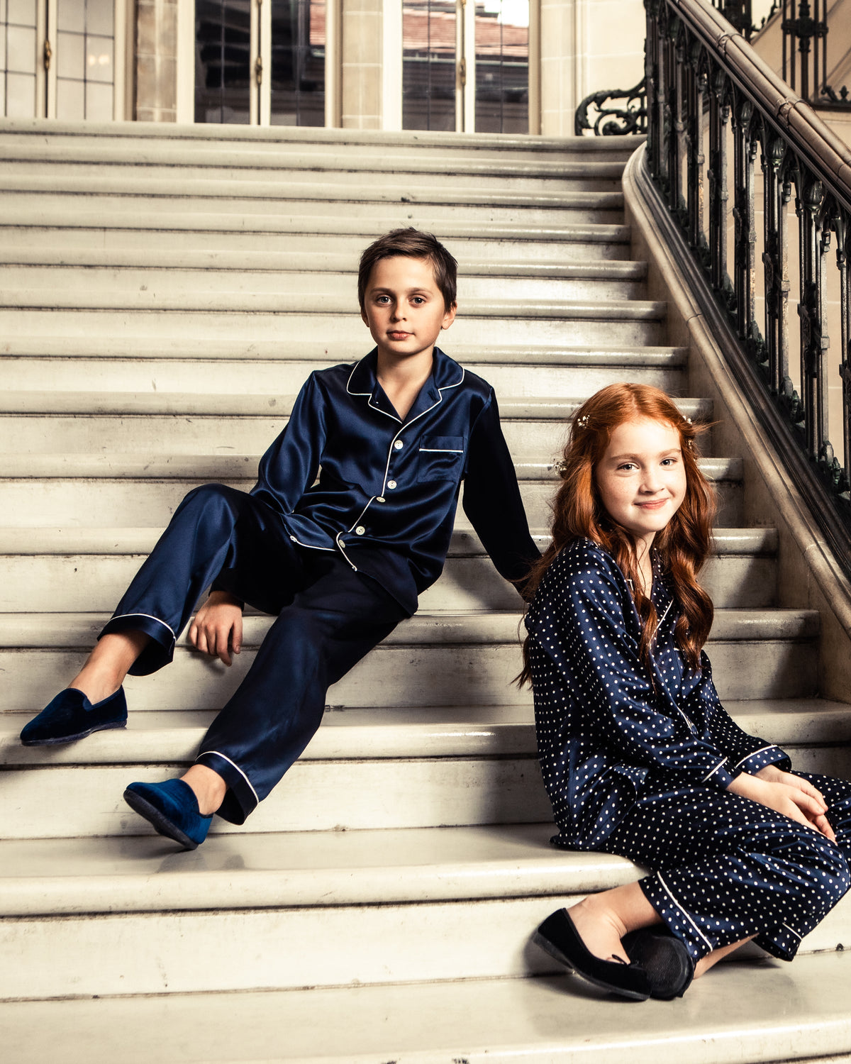 Two children in navy pajamas sit on a staircase; one wears The Gable navy velvet slipper