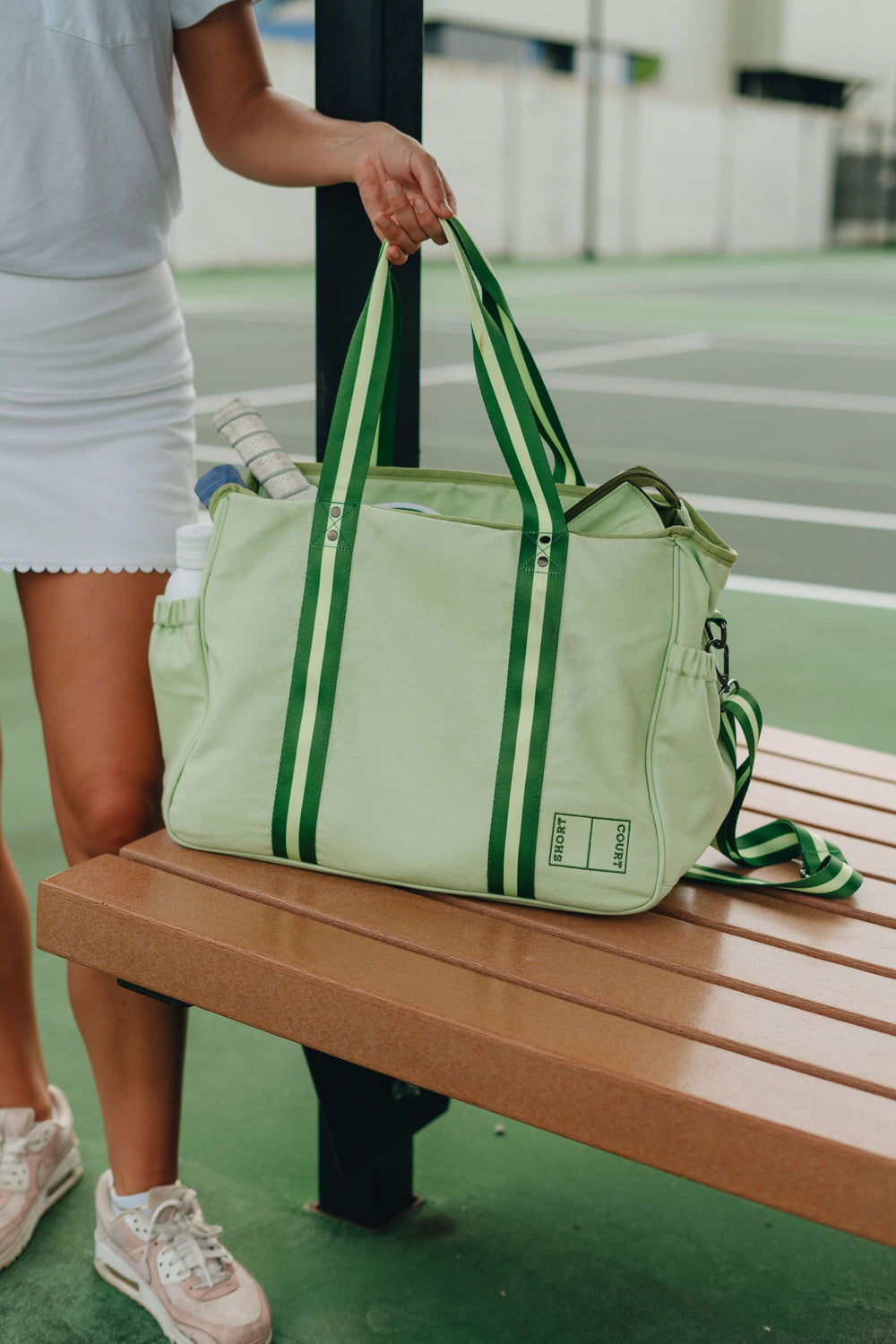 Side view of Short Court Tote with pale green canvas, dark green striped handles, exterior paddle pocket on tennis court bench