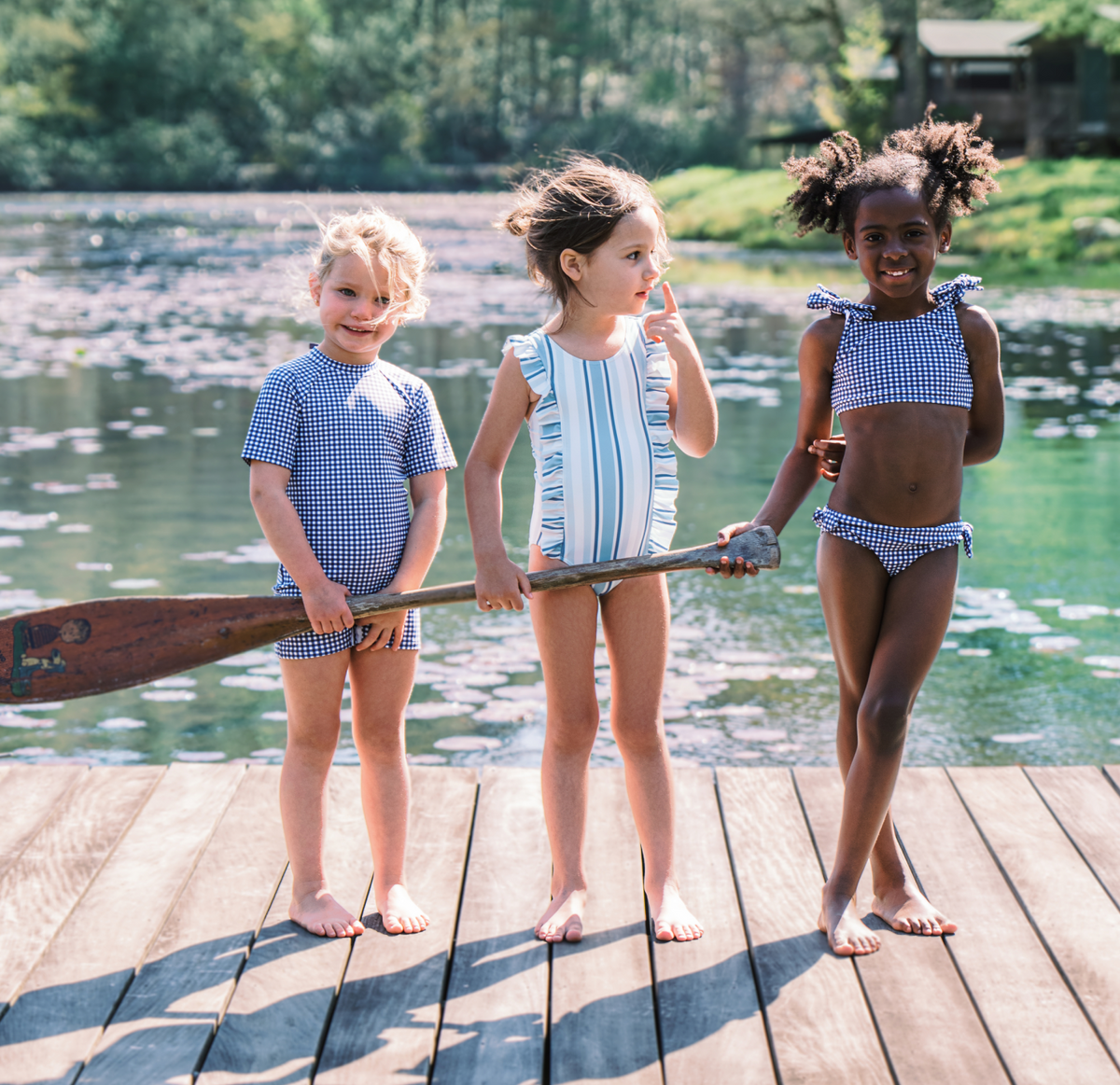 Three girls stand on a wooden dock by a lake; the girl on the right wears a navy gingham tie-knot bikini and holds a paddle