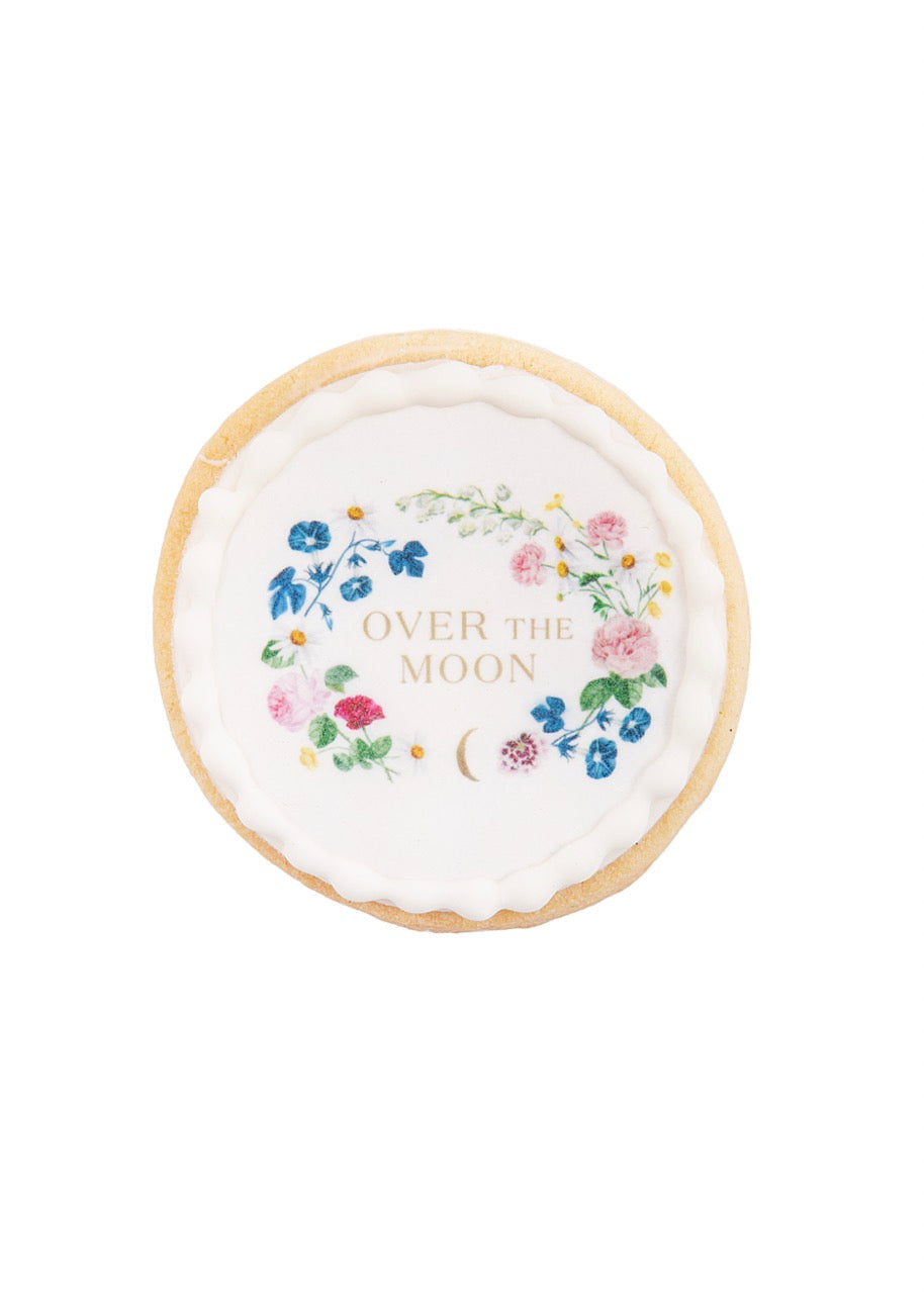 Top-down view of a crest sugar cookie with floral design and gold OVER THE MOON text on white royal icing on a white background