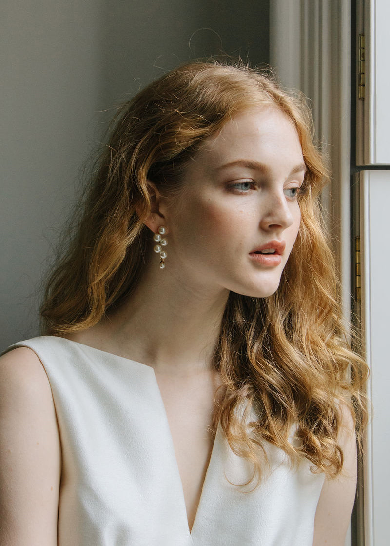 Side view of Marcella Earrings showcasing dangling gold design with a softly lit window background