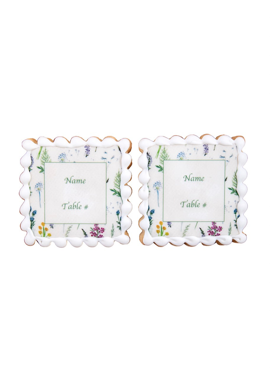 Front view of floral multicolor name plate sugar cookies showing scalloped white icing and floral center labeled ‘Name’ and ‘Table #’ on white background