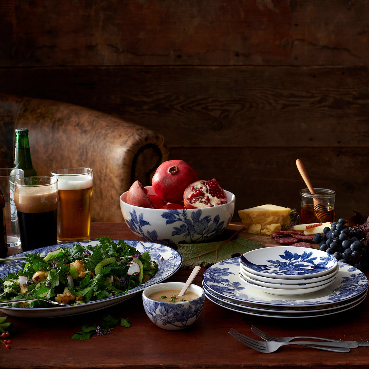 Blue Arbor dinnerware arranged on a rustic wooden table with salad on a large oval platter, stacked plates, fruit bowl, cheese, and drinks