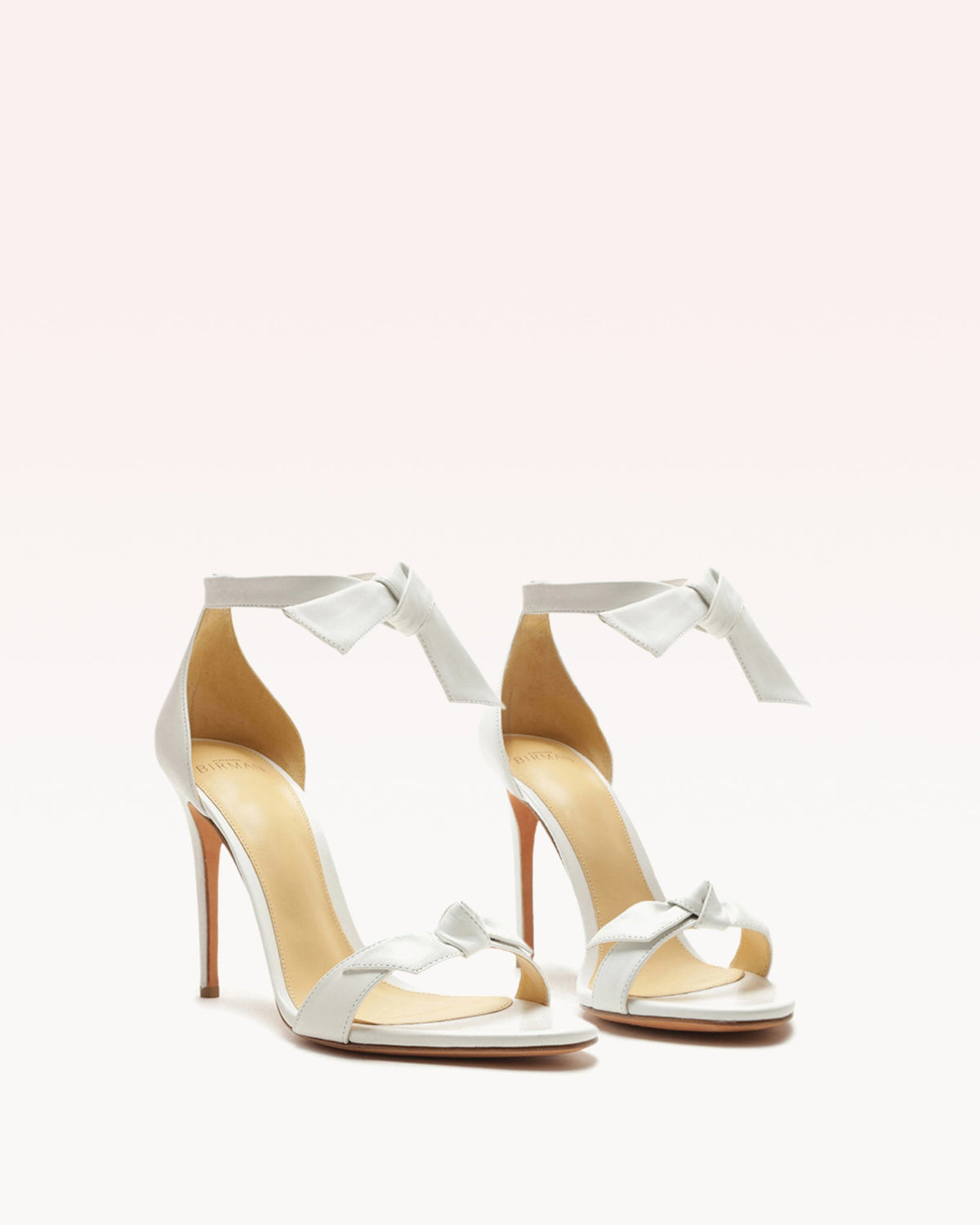 Front three-quarter view of Clarita 100 in White Leather sandals showcasing two bow accents and stiletto heels against a light background