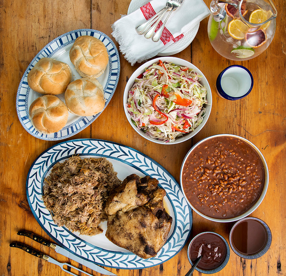 Overhead of Carolina-style BBQ kit with pulled pork, roasted chicken, baked beans, coleslaw, rolls, and BBQ sauces