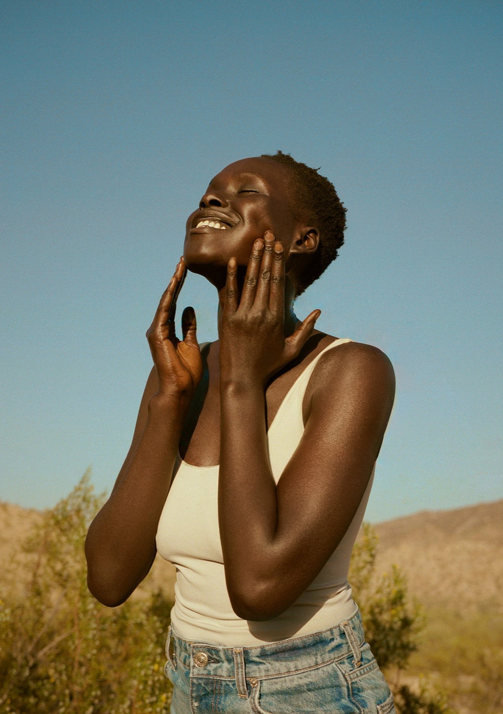 Woman smiling with eyes closed massaging her face with both hands outdoors against a clear blue sky