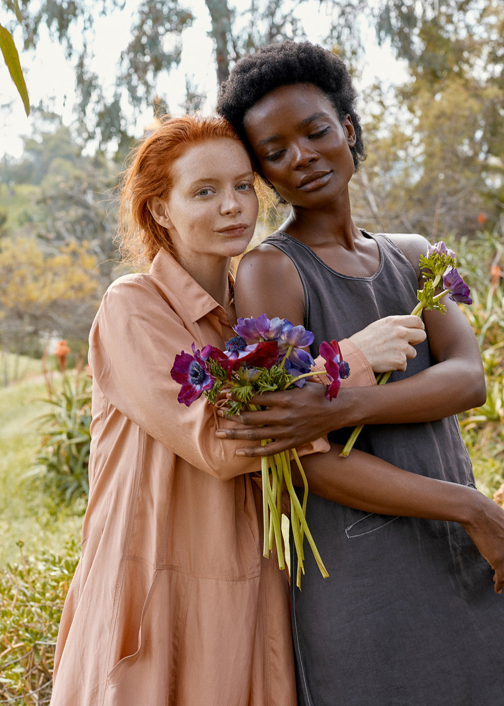 Two models embrace while holding purple and magenta flowers in a sunlit outdoor garden setting