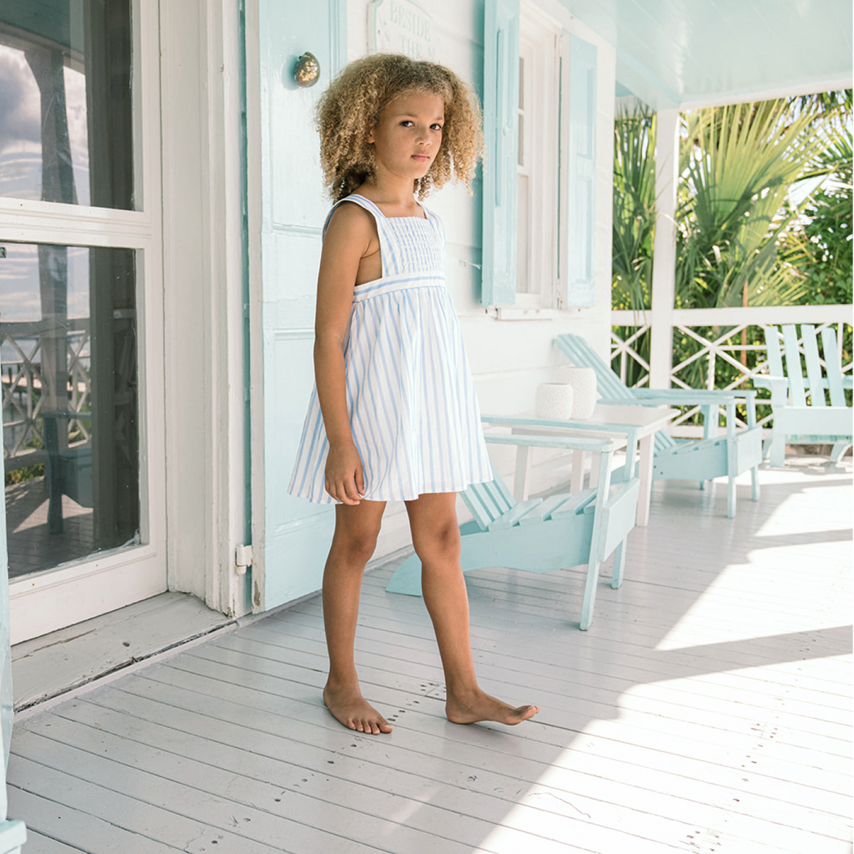 Front view of Girls Bahamian Blue Stripe Smocked Bib Dress in a relaxed fit on a child, showing smocked panel on a sunny porch